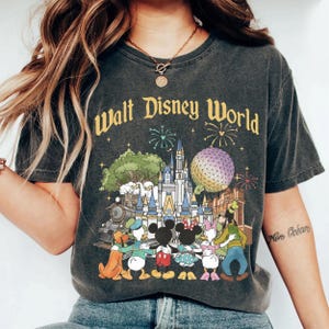 May include: Dark gray t-shirt featuring a colorful graphic of the Walt Disney World logo and iconic park imagery. The design includes a castle, characters, fireworks, and the text "Walt Disney World".