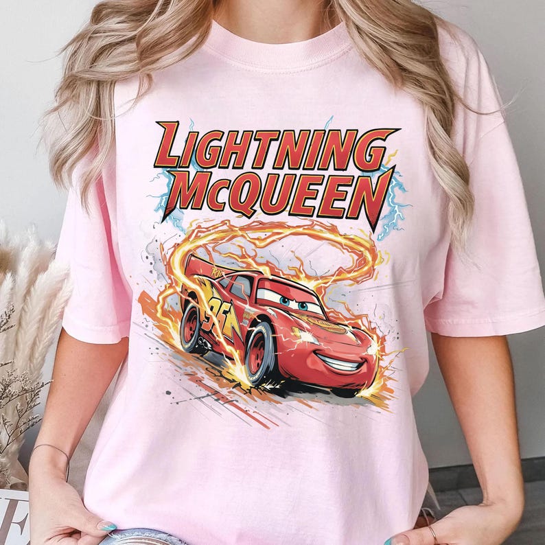 Może przedstawiać: Jasnor&oacute;żowa koszulka z grafiką Zygzaka McQueena, czerwonego samochodu wyścigowego z animowanego filmu Auta. Tekst "LIGHTNING McQUEEN" znajduje się nad samochodem w kolorze czerwonym i ż&oacute;łtym. Samoch&oacute;d otoczony jest płomieniami.