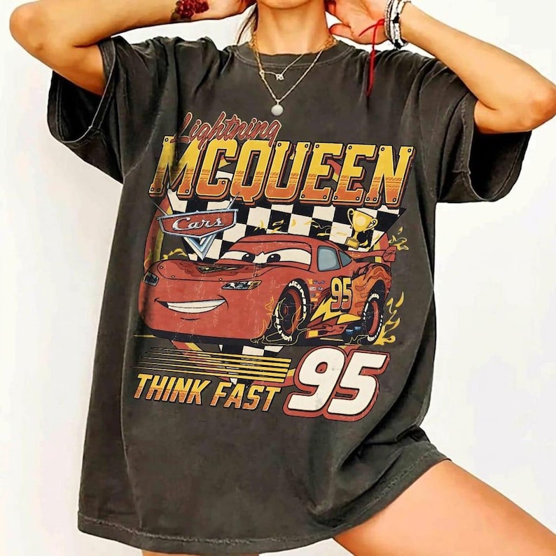 May include: Dark gray t-shirt featuring a graphic of Lightning McQueen from the movie Cars. The design includes the text "Lightning McQueen," "Cars," "Think Fast," and the number "95" in a vintage style.