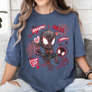 Op de afbeelding: Blauw t-shirt met een cartoonillustratie van een superheld in een zwart en rood pak. Het ontwerp bevat de woorden "AMAZING", "MILES", "THWIP", "WEB-HEAD" en "GO-WEBS-GO!" in het rood, samen met spinnenwebben en harten.