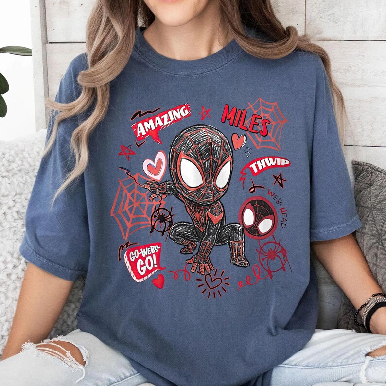 Op de afbeelding: Blauw t-shirt met een cartoonillustratie van een superheld in een zwart en rood pak. Het ontwerp bevat de woorden "AMAZING", "MILES", "THWIP", "WEB-HEAD" en "GO-WEBS-GO!" in het rood, samen met spinnenwebben en harten.