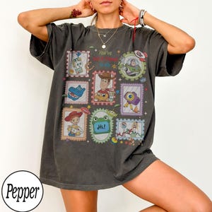 May include: A charcoal gray short-sleeve t-shirt featuring a graphic print of various Toy Story characters arranged in rectangular and circular frames. The text "You've Got A Friend In Me" and "Hi!" are printed among the colorful character illustrations.