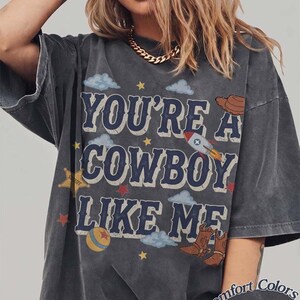 May include: A charcoal gray short-sleeve t-shirt featuring the text "YOU'RE A COWBOY LIKE ME" in blue block letters. The design includes illustrations of a rocket, a cowboy hat, a sheriff star, a ball, and small stars and clouds.