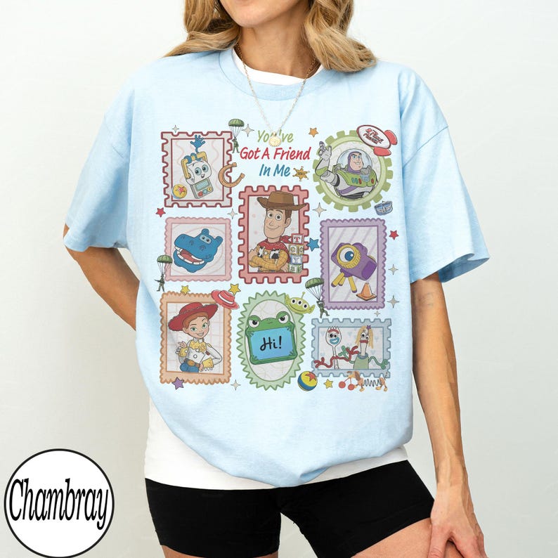 May include: A light blue t-shirt featuring a graphic print of various Toy Story characters arranged in postage stamp-style frames. The design includes Woody, Buzz Lightyear, Jessie, and Forky, with the text "You've Got A Friend In Me" and "Hi!" printed among the colorful illustrations.