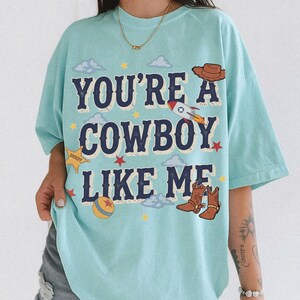 May include: A light blue t-shirt featuring the dark blue text "YOU'RE A COWBOY LIKE ME" surrounded by graphic illustrations of a brown cowboy hat, a rocket ship, a yellow sheriff star badge, a colorful ball, and brown cowboy boots with clouds.