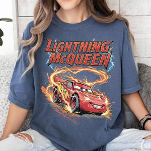 Może przedstawiać: Niebieska koszulka z grafiką Zygzaka McQueena, czerwonego samochodu wyścigowego z numerem 95, otoczonego płomieniami i błyskawicami. Tekst "Lightning McQueen" jest wyświetlany nad samochodem w kolorze czerwonym i ż&oacute;łtym.