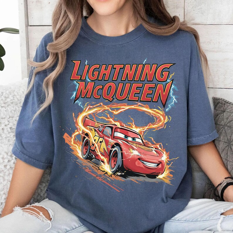 Może przedstawiać: Niebieska koszulka z grafiką Zygzaka McQueena, czerwonego samochodu wyścigowego z numerem 95, otoczonego płomieniami i błyskawicami. Tekst "Lightning McQueen" jest wyświetlany nad samochodem w kolorze czerwonym i ż&oacute;łtym.