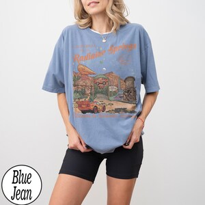 Pu&ograve; includere: Una maglietta blu con una grafica di Radiator Springs, con il testo "California Radiator Springs". Il design include auto e paesaggi desertici. La maglietta &egrave; abbinata a pantaloncini neri.