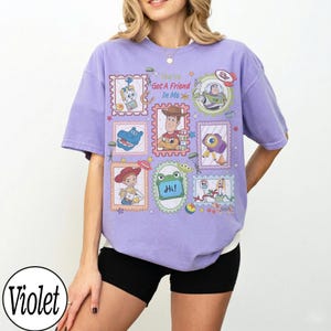 May include: A light violet short-sleeve t-shirt featuring a graphic print of various Toy Story characters in postage stamp-style frames. The text "You've Got A Friend In Me" is printed in the center, along with a small green frog holding a sign saying "Hi!"