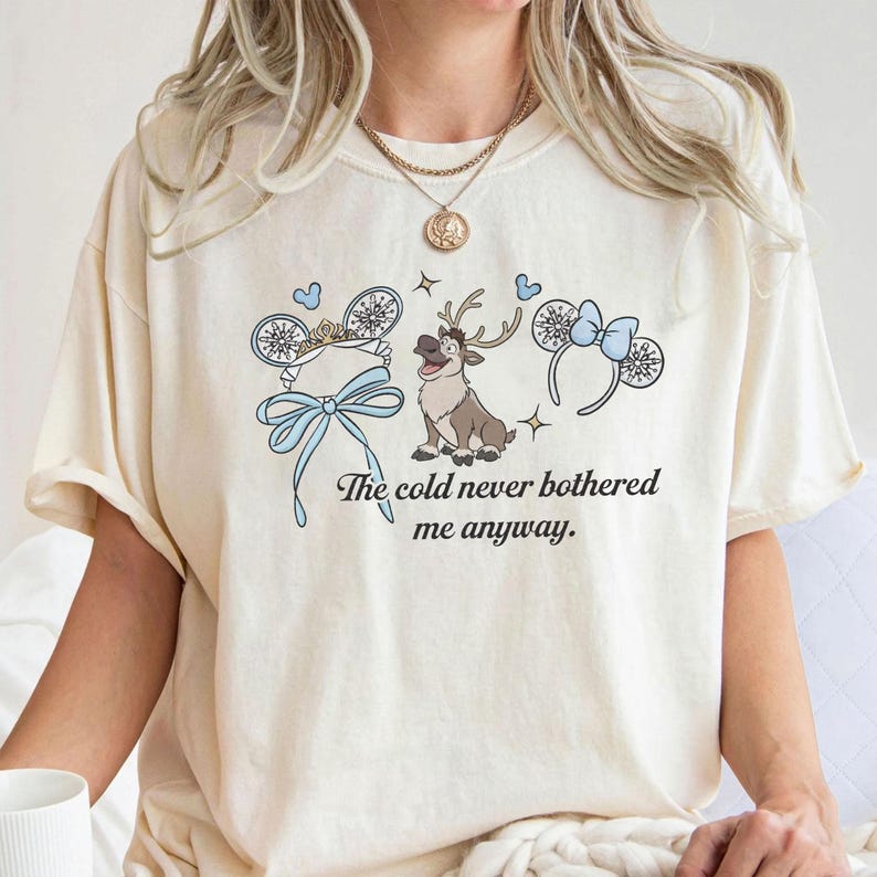 May include: Cream-coloured t-shirt with a cartoon reindeer and mouse ear headbands with blue bows and snowflakes. The text "The cold never bothered me anyway" is printed below the graphic.