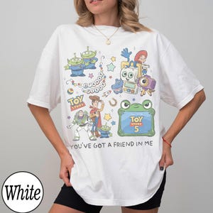Puede incluir: Una camiseta blanca con un estampado gr&aacute;fico colorido de personajes de Toy Story, incluyendo a Woody, Buzz Lightyear y los Aliens. El dise&ntilde;o incluye el texto Disney Pixar, Toy Story 5 y la frase You've Got A Friend In Me.
