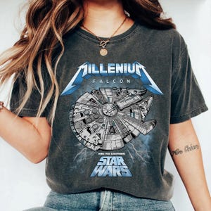 Puede incluir: Una camiseta de manga corta en color gris carbón con un gráfico de la nave espacial Millennium Falcon. El diseño incluye el texto "MILLENIUM FALCON" y "RIDE THE LIGHTNING STAR WARS" en letras azules, acentuado con rayos blancos.