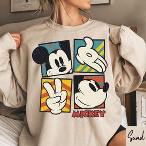 Puede incluir: Sudadera beige con un dise&ntilde;o de cuatro paneles con im&aacute;genes de Mickey Mouse. Cada panel muestra un elemento diferente: la cara de Mickey, una mano enguantada, un signo de la paz y otra cara de Mickey. La palabra "MICKEY" est&aacute; impresa en rojo.