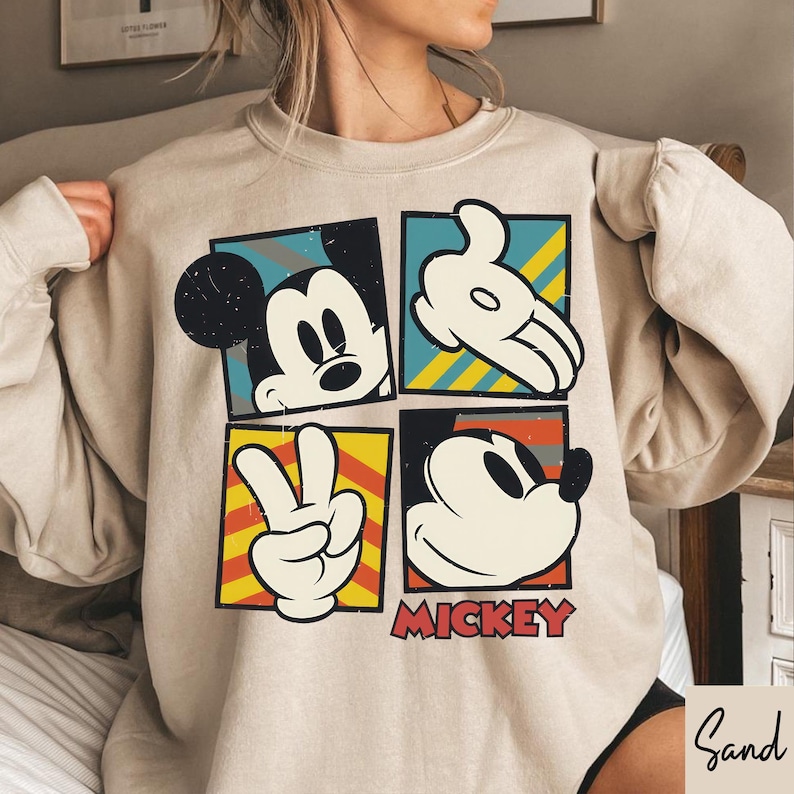 Puede incluir: Sudadera beige con un dise&ntilde;o de cuatro paneles con im&aacute;genes de Mickey Mouse. Cada panel muestra un elemento diferente: la cara de Mickey, una mano enguantada, un signo de la paz y otra cara de Mickey. La palabra "MICKEY" est&aacute; impresa en rojo.