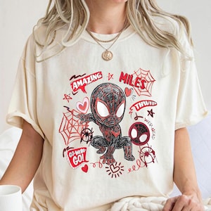 Op de afbeelding: Cr&egrave;mekleurig T-shirt met een cartoonillustratie van Miles Morales in een Spider-Man-pak. Het ontwerp bevat rode accenten, spinnenwebben, harten en de woorden "Amazing", "Miles", "Thwip" en "Go-Webs-Go!"
