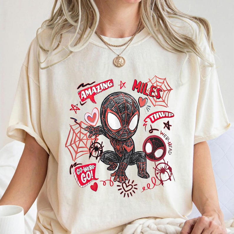 Op de afbeelding: Cr&egrave;mekleurig T-shirt met een cartoonillustratie van Miles Morales in een Spider-Man-pak. Het ontwerp bevat rode accenten, spinnenwebben, harten en de woorden "Amazing", "Miles", "Thwip" en "Go-Webs-Go!"