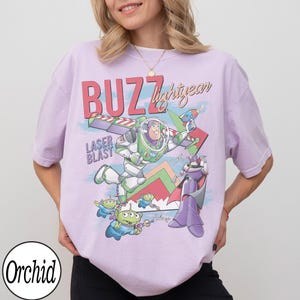 May include: A light purple t-shirt featuring a graphic of Buzz Lightyear from Toy Story. The design includes the words "BUZZ Lightyear" and "LASER BLAST", along with images of aliens and Zurg.