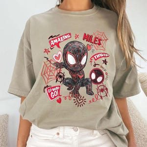 Op de afbeelding: Lichtbruin t-shirt met een cartoonontwerp van Spider-Man. De afbeelding bevat de rode woorden "AMAZING MILES" en "GO-WEBS-GO!", evenals spinnenwebben, harten en sterren. Het ontwerp is speels.