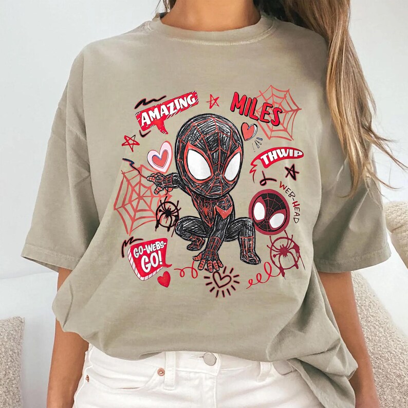 Op de afbeelding: Lichtbruin t-shirt met een cartoonontwerp van Spider-Man. De afbeelding bevat de rode woorden "AMAZING MILES" en "GO-WEBS-GO!", evenals spinnenwebben, harten en sterren. Het ontwerp is speels.