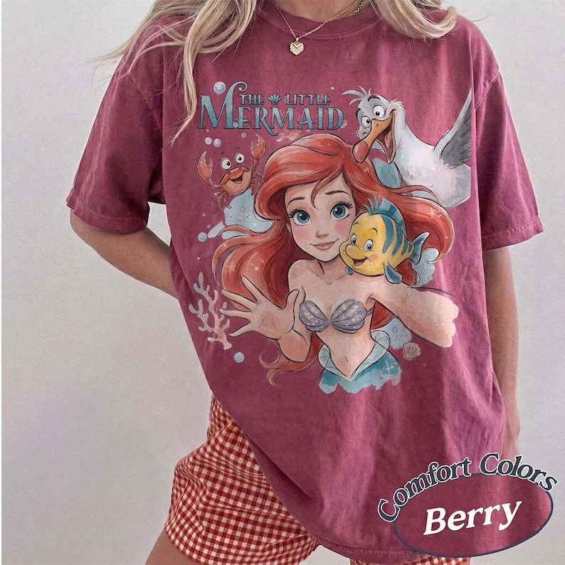 May include: A berry-colored short-sleeve t-shirt featuring a graphic print of Ariel from The Little Mermaid, along with Flounder, Sebastian, and Scuttle. The text The Little Mermaid is printed above the characters in a blue and white font.