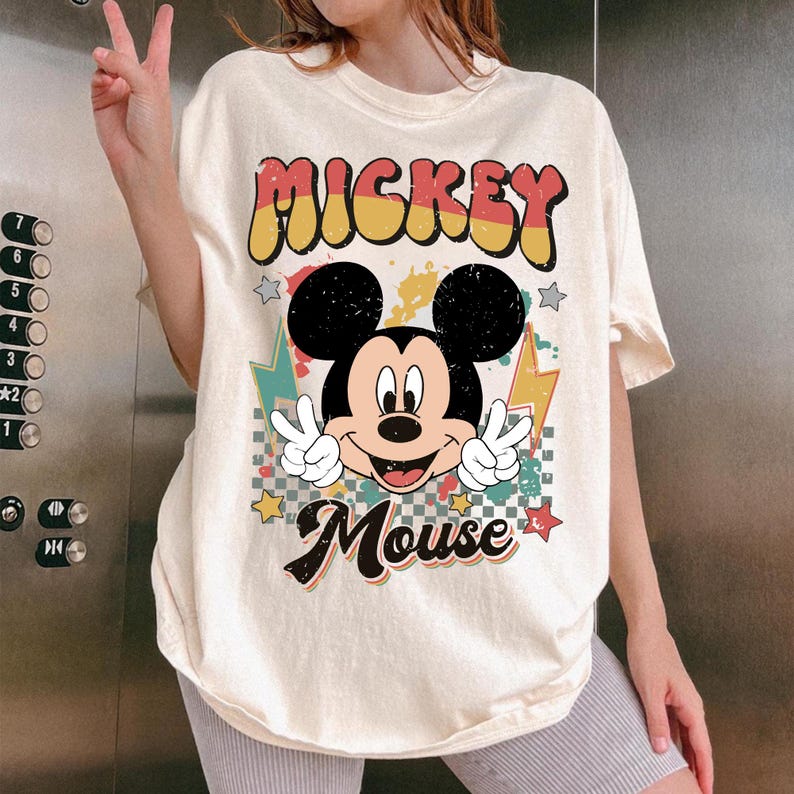 May include: Cream-colored t-shirt featuring a retro Mickey Mouse graphic. The design includes the words "Mickey Mouse" and "Mickey" in a vintage font, along with Mickey Mouse making a peace sign. The shirt has a relaxed fit.