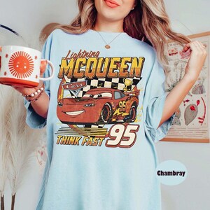 May include: Light blue t-shirt featuring Lightning McQueen from the Cars movie. The graphic includes the text "Lightning McQueen", "Cars", "Think Fast", and the number "95". A red and orange mug with a sun design is held in the hand.