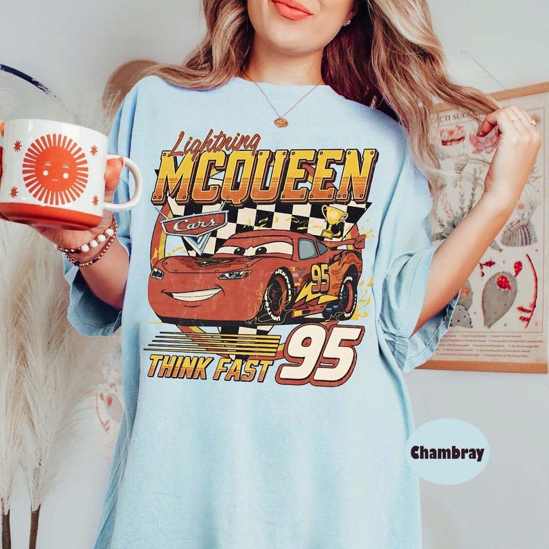 May include: Light blue t-shirt featuring Lightning McQueen from the Cars movie. The graphic includes the text "Lightning McQueen", "Cars", "Think Fast", and the number "95". A red and orange mug with a sun design is held in the hand.