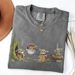 May include: A charcoal gray crew neck t-shirt featuring a graphic print of four small green creatures with large ears and tan robes. One creature is near a tree, one is in a floating pod, one stands, and one sits on a log. The shirt has a Comfort Colors label.