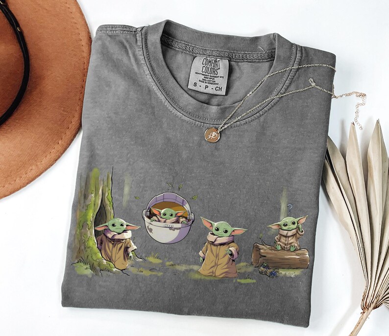 May include: A charcoal gray crew neck t-shirt featuring a graphic print of four small green creatures with large ears and tan robes. One creature is near a tree, one is in a floating pod, one stands, and one sits on a log. The shirt has a Comfort Colors label.
