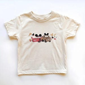May include: Off-white toddler t-shirt with a graphic of two cartoon cars wearing Mickey Mouse ears. A red car and a brown truck are surrounded by food icons. The shirt is made of a soft, comfortable material.