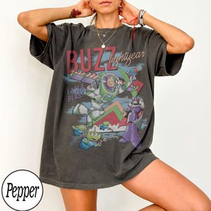 May include: Dark gray t-shirt featuring a graphic of Buzz Lightyear with the text "BUZZ lightyear" and "LASER BLAST". The design includes cartoon aliens and a purple villain. The shirt has short sleeves and a relaxed fit.