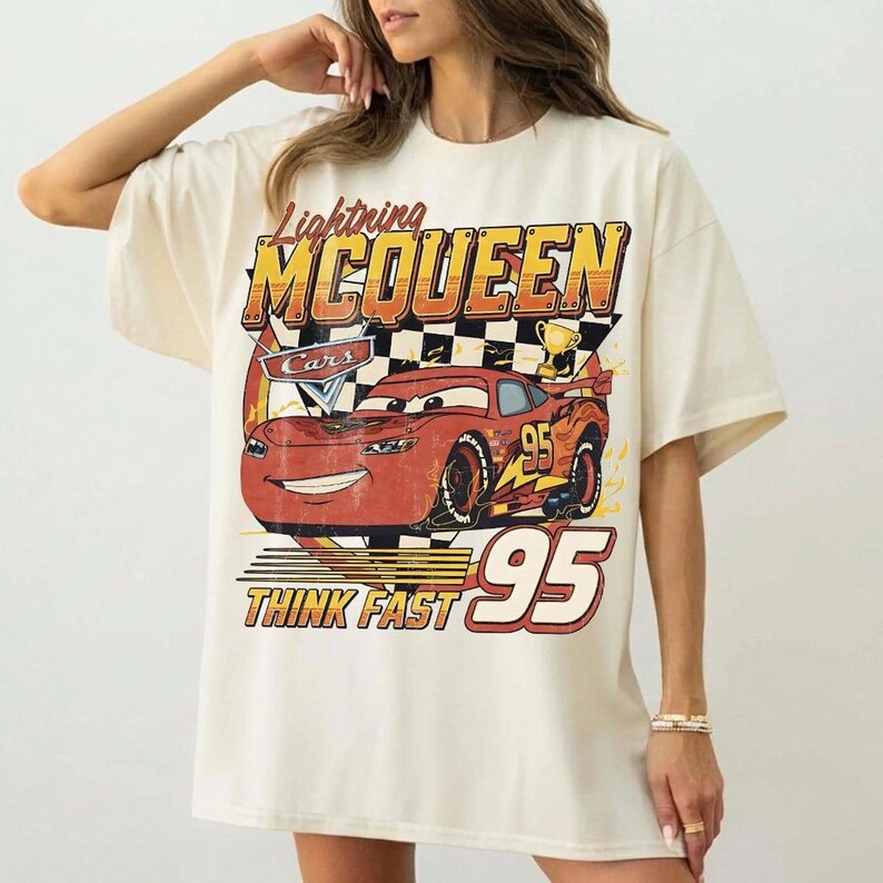 May include: Cream-colored t-shirt featuring a graphic of Lightning McQueen from the Cars movie. The design includes the text "Lightning McQueen", "Cars", "Think Fast", and the number "95". The shirt has short sleeves and a relaxed fit.