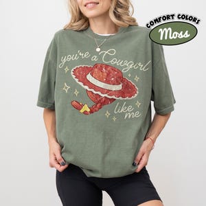 May include: A moss green short-sleeve t-shirt featuring a graphic of a red cowgirl hat with a braided ponytail and the text you're a Cowgirl like me in white cursive script, accented by small yellow star shapes across the front of the garment.