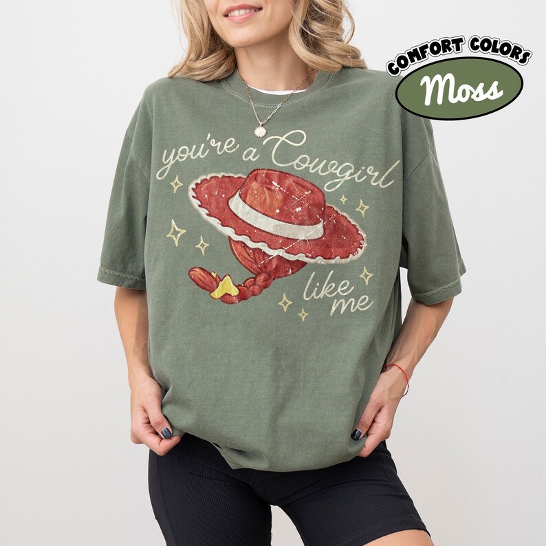 May include: A moss green short-sleeve t-shirt featuring a graphic of a red cowgirl hat with a braided ponytail and the text you're a Cowgirl like me in white cursive script, accented by small yellow star shapes across the front of the garment.