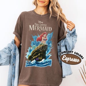 May include: A dark brown short-sleeve t-shirt featuring a graphic of Ariel from The Little Mermaid sitting on a rock amidst blue ocean waves. The text Disney The Little Mermaid is printed above the character. A Comfort Colors Espresso label is visible.