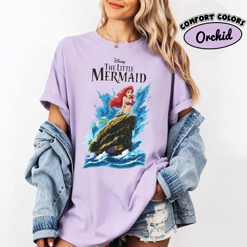 May include: A light purple t-shirt featuring a graphic of Ariel from Disney's The Little Mermaid sitting on a rock amidst blue ocean waves. The text reads Disney The Little Mermaid. The shirt is worn with a blue denim jacket and denim shorts.