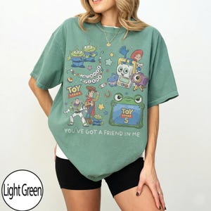Puede incluir: Una camiseta de manga corta de color verde claro con un estampado gr&aacute;fico de personajes de Toy Story, incluyendo a Woody, Buzz Lightyear y alien&iacute;genas. El dise&ntilde;o incluye el texto Disney Pixar, Toy Story, Toy Story 5 y You've Got A Friend In Me.
