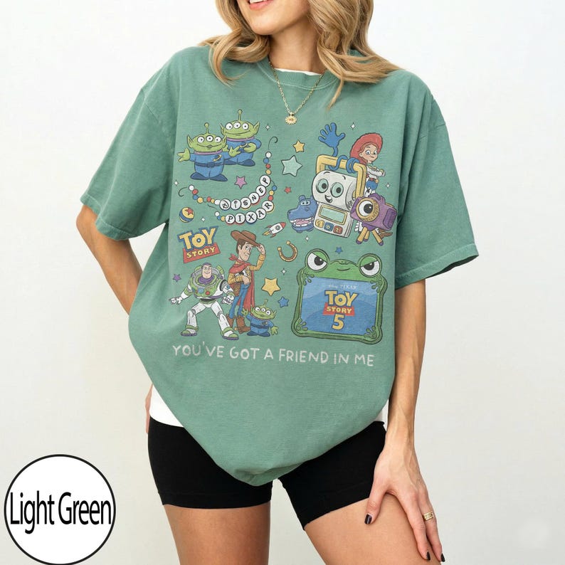 Puede incluir: Una camiseta de manga corta de color verde claro con un estampado gr&aacute;fico de personajes de Toy Story, incluyendo a Woody, Buzz Lightyear y alien&iacute;genas. El dise&ntilde;o incluye el texto Disney Pixar, Toy Story, Toy Story 5 y You've Got A Friend In Me.