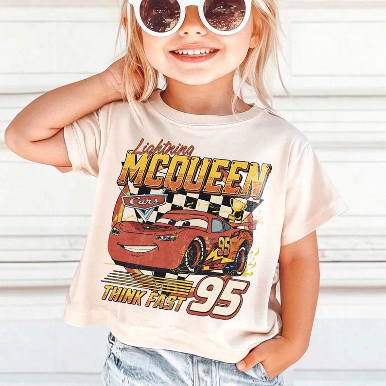 May include: A beige t-shirt featuring a graphic of Lightning McQueen from the Cars movie. The design includes the text "Lightning McQueen", "Cars", "Think Fast", and the number "95". The shirt has short sleeves.