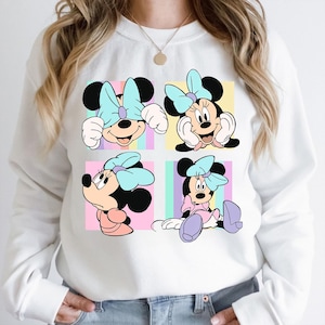 May include: White sweatshirt with four Minnie Mouse designs, each with a light blue bow, set against pastel backgrounds. The sweatshirt has a crew neck and long sleeves.