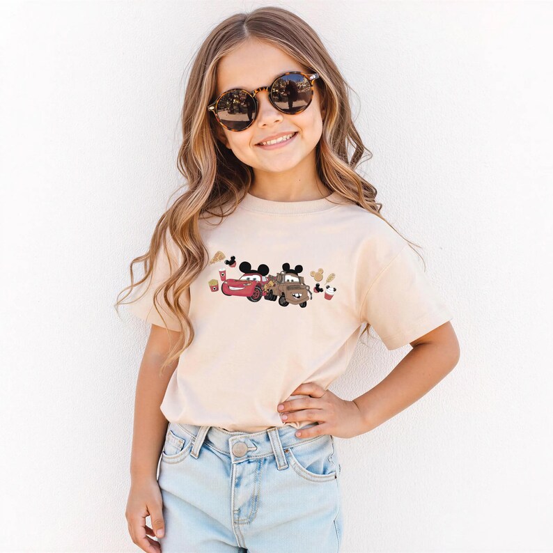 May include: A beige short-sleeved t-shirt with a colourful graphic of cartoon cars and Mickey Mouse characters. The design includes red and brown cars, and Mickey Mouse ears. The shirt is worn by a young girl wearing sunglasses.