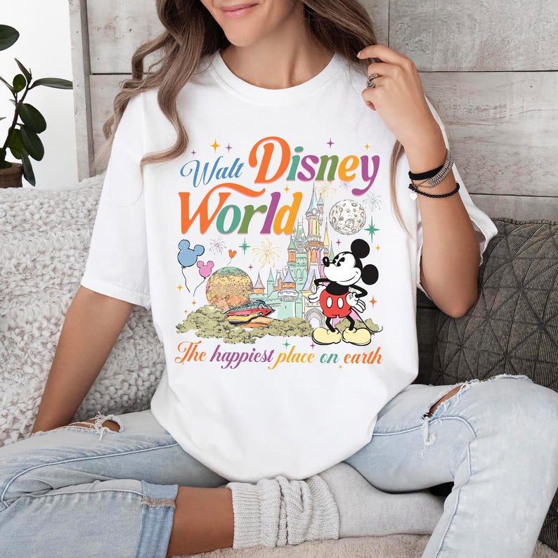 以下が含まれることがあります： ウォルト・ディズニー・ワールドのカラフルなグラフィックが特徴の白いTシャツ。デザインにはミッキーマウス、お城、そして「The happiest place on earth」というフレーズが含まれています。テキストはマルチカラーです。