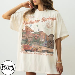Pu&ograve; includere: T-shirt color avorio con grafica di Radiator Springs, con testo "California Est. 2012" e "Welcome to Radiator Springs." Il design include un'auto rossa, una torre dell'acqua e un paesaggio desertico. La maglietta &egrave; oversize.
