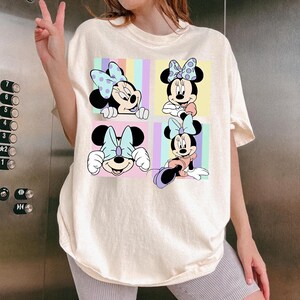 May include: Cream-colored t-shirt featuring a four-panel graphic of Minnie Mouse. Each panel showcases a different pose of Minnie, with her signature bow and various pastel backgrounds. The shirt has short sleeves and a relaxed fit.