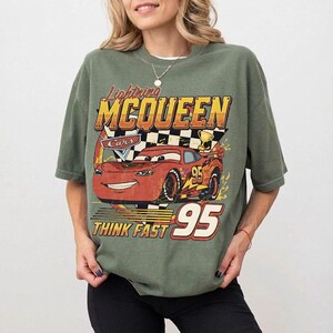 May include: Olive green t-shirt featuring a graphic of Lightning McQueen from the Cars movie. The design includes the text "Lightning McQueen", "Cars", "Think Fast", and the number "95" in a vintage style.