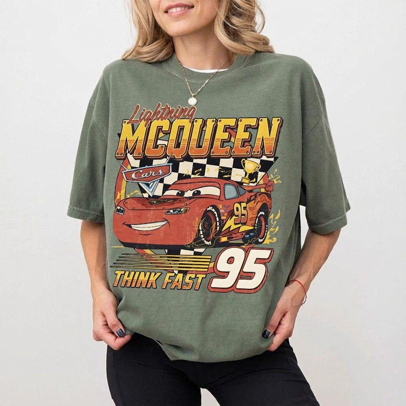 May include: Olive green t-shirt featuring a graphic of Lightning McQueen from the Cars movie. The design includes the text "Lightning McQueen", "Cars", "Think Fast", and the number "95" in a vintage style.