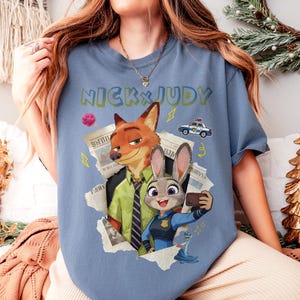 May include: A blue t-shirt featuring the text "NICK & JUDY" in green and yellow. The graphic includes cartoon images of a fox and a rabbit, along with a police car and other design elements. The fox is wearing a green shirt and tie, and the rabbit is wearing a police uniform.
