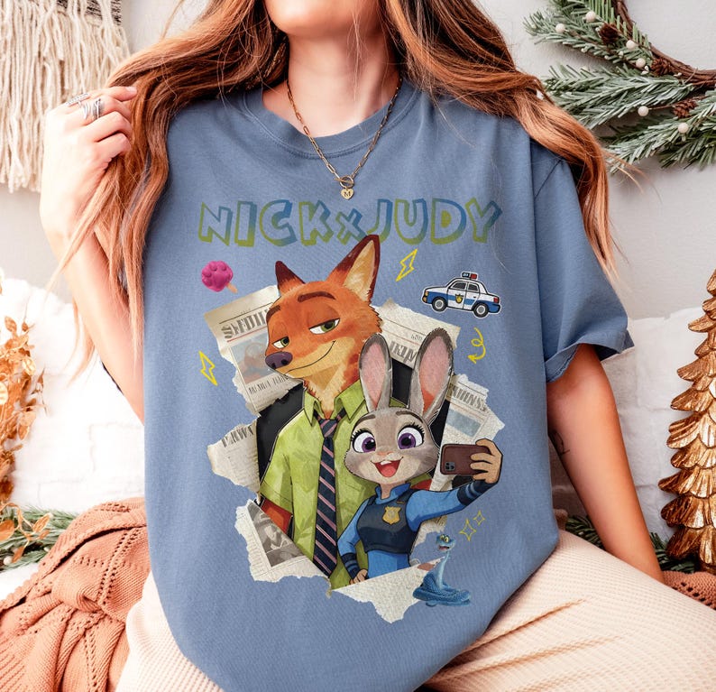 May include: A blue t-shirt featuring the text "NICK & JUDY" in green and yellow. The graphic includes cartoon images of a fox and a rabbit, along with a police car and other design elements. The fox is wearing a green shirt and tie, and the rabbit is wearing a police uniform.