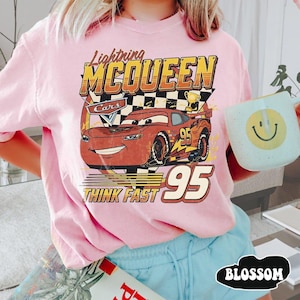 May include: Pink t-shirt featuring a graphic of Lightning McQueen from the Cars movie. The design includes the text "Lightning McQueen", "Cars", "95", and "Think Fast". The shirt has short sleeves and a crew neck.