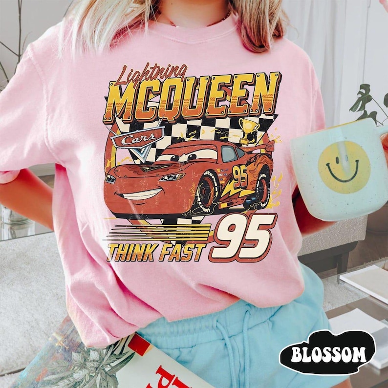 May include: Pink t-shirt featuring a graphic of Lightning McQueen from the Cars movie. The design includes the text "Lightning McQueen", "Cars", "95", and "Think Fast". The shirt has short sleeves and a crew neck.